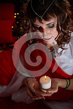 Beautiful arabian girl with candles in red room full of rich fabrics and carpets in sultan harem. Photo shoot of woman