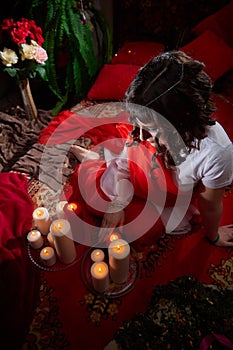 Beautiful arabian girl with candles in red room full of rich fabrics and carpets in sultan harem. Photo shoot of woman