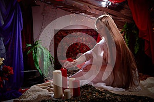 Beautiful arabian girl with candles in red room full of rich fabrics and carpets in sultan harem. Photo shoot of woman