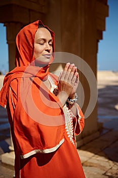 Beautiful Arabian Muslim woman in bright orange authentic dress and her head covered in hijab, praying with her hands up