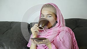 Beautiful arabian woman in pink hijab sitting on a sofa in a cafe and drinks cofee