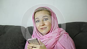 Beautiful arabian woman in pink hijab sitting on a sofa in a cafe and sends a text message on cell phone