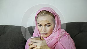 Beautiful arabian woman in pink hijab sitting on a sofa in a cafe and sends a text message on cell phone