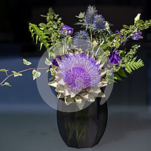 Purple artichoke flower with thistles in a small black glass vase against dark background.