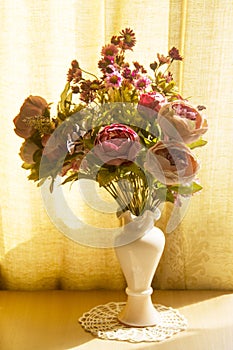 Beautiful artificial flowers of peonies and chrysanthemums in a delicate pink vase on a warm ocher yellow background