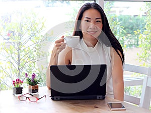 Asian woman holding cup of coffee smiling and looking to the camera.computer and glass and flower pot on table