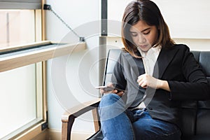 A beautiful asian business woman sitting on sofa holding smart phone and checking her suit for meeting today.