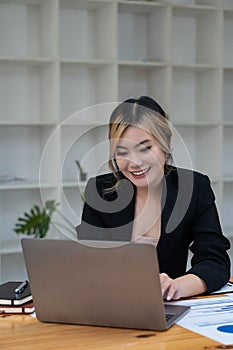 Beautiful Asian businesswoman analyzing chart using calculator, laptop at the office with papers and holding pen, sitting on the c