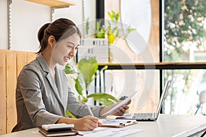 Beautiful Asian businesswoman analyzing chart using calculator, laptop at the office with papers and holding pen, sitting on the c