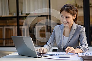 Beautiful Asian businesswoman analyzing chart using calculator, laptop at the office with papers and holding pen, sitting on the c