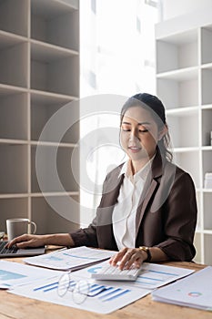 Beautiful Asian businesswoman analyzing charts using calculator on laptop at office with documents