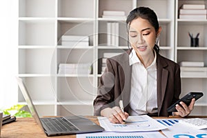 Beautiful Asian businesswoman analyzing charts using calculator on laptop at office with documents