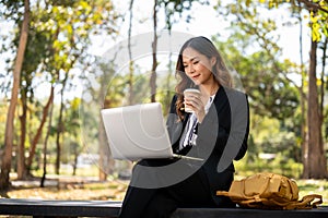 A beautiful Asian businesswoman is sitting on a bench in a city park, and working on her laptop