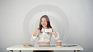 A beautiful Asian businesswoman sits at her desk, showing her thumb up and holding a smartphone