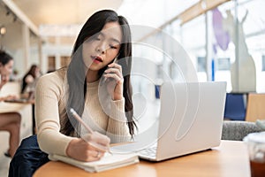 A beautiful Asian businesswoman is taking notes in her notebook and having a serious phone call