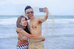 beautiful Asian Chinese couple taking selfie photo with mobile phone camera smiling joyful having fun on the beach