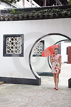 Beautiful Asian Chinese woman in traditional chi-pao cheongsam hold red parasol in a garden stand under arched door