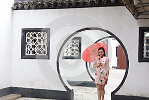 Beautiful Asian Chinese woman in traditional chi-pao cheongsam hold red parasol in a garden stand under arched door