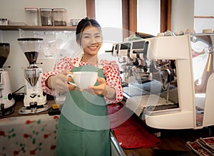 Beautiful asian coffee girl wearing apron lodge a coffee cup to the camera with smile in coffee shop