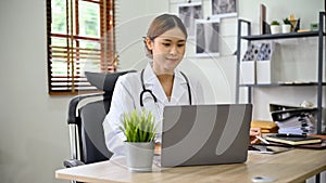 Beautiful Asian female doctor working at her office desk, using laptop computer