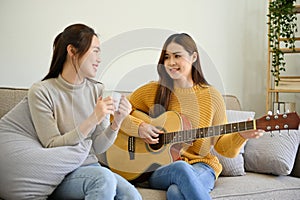 Beautiful Asian female playing guitar and her friend singing, having fun time together at home