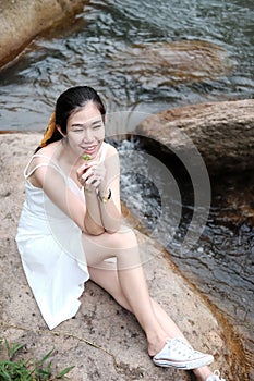 Beautiful asian female in white dress is hand holding grass flower on the rock and stream in forest
