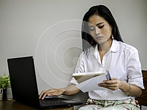 Beautiful asian freelancer looking note book, typing on labtop keyboard concentrate her work