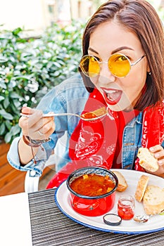 beautiful asian girl eating hungarian goulash soup from a decorative red casserole in outdoor restaurant