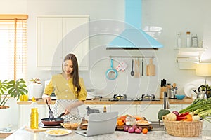 A beautiful Asian girl makes breakfast by looking at how to make it with her laptop computer with a happy face. At her own house