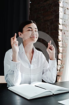 Beautiful asian girl sitting at a table for an interview and crossing her fingers in the hope of closing her eyes