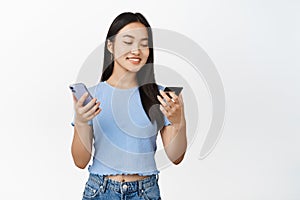 Beautiful asian girl smiling, using credit card and mobile phone, order on smartphone, paying contactless, white