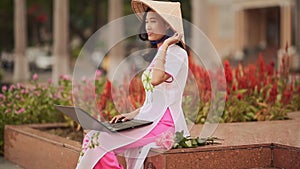 The beautiful Asian girl speaks on a laptop on the street and a beautiful national dress Ao Dai.