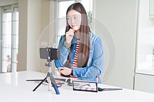 Beautiful Asian influencer woman recording make up tutorial using smartphone camera with hand on chin thinking about question,