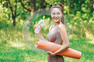 Beautiful asian lady holding fitness mat and bottle of water, preparing for yoga practise in a forest meadow.