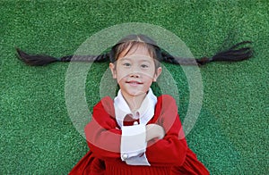 Beautiful Asian little girl in red dress cross one`s arm and lying on green grass background in the winter season and happy new