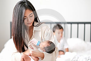 Beautiful Asian mother give milk to her newborn baby via bottle with her little boy as background on bed