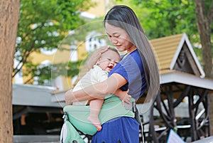 Beautiful Asian mother hugging her infant on ergonomic baby carrier in the garden