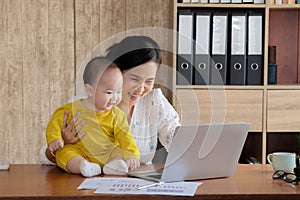 Beautiful Asian mother spent time with curiosity toddler baby boy talking, playing on workplace home, adorable naughty son happy