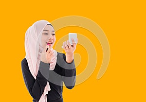 Beautiful Asian muslim woman in hijab standing holding mirror and applying pink lipstick on her lips isolated on yellow background