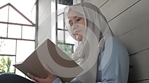Beautiful Asian muslim woman reading book while sitting outside of her home, girl enjoys her time by doing leisure activity