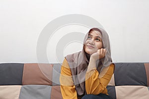 Beautiful Asian muslim woman thinking something good while sitting on sofa, girl with contemplation expression, looking up and