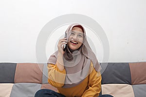 Beautiful Asian muslim woman using phone while sitting on sofa, girl talking on smartphone and smiling