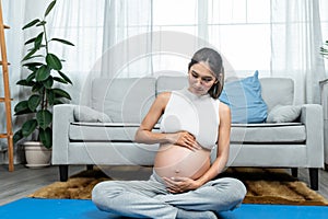 Beautiful Asian pregnant woman, big belly, sitting on blue exercise mat, she sat down and gently stroked growing belly, stimulate