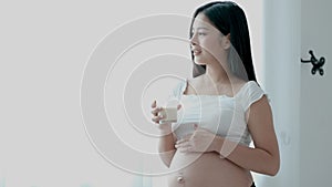 Beautiful Asian pregnant woman drink milk in front of glass window and feel happy then smile and also look to her belly
