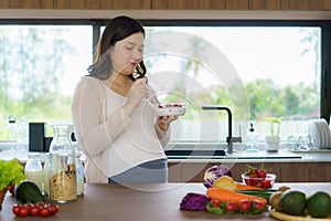 Beautiful Asian pregnant woman eating with lust cereals on breakfast in kitchen at home, enjoying meal. Yummy food for pregnant