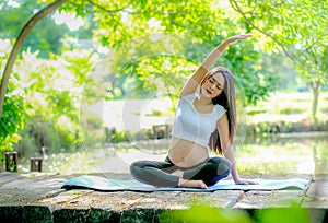 Beautiful Asian pregnant woman exercise with yoga action by sit on wood bridge near the river in the garden with morning light