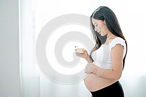 Beautiful Asian pregnant woman hold glass of milk and look to her belly in front white curtain and she look happy by smiling.