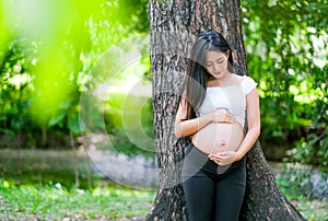 Beautiful Asian pregnant woman lean on the tree, look at and touch her belly in green garden with morning light