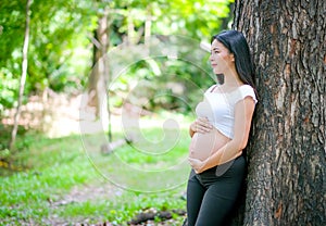 Beautiful Asian pregnant woman smile, lean at the tree and touch her belly with look at the side among green garden with morning