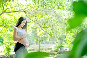 Beautiful Asian pregnant women look at and touch her belly with green background of garden and also leaf as foreground